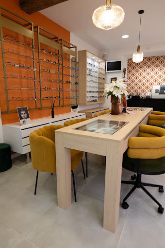 L’intérieur de la boutique : mur de montures, table en bois clair, fauteuils moutarde et papier peint géométrique orange