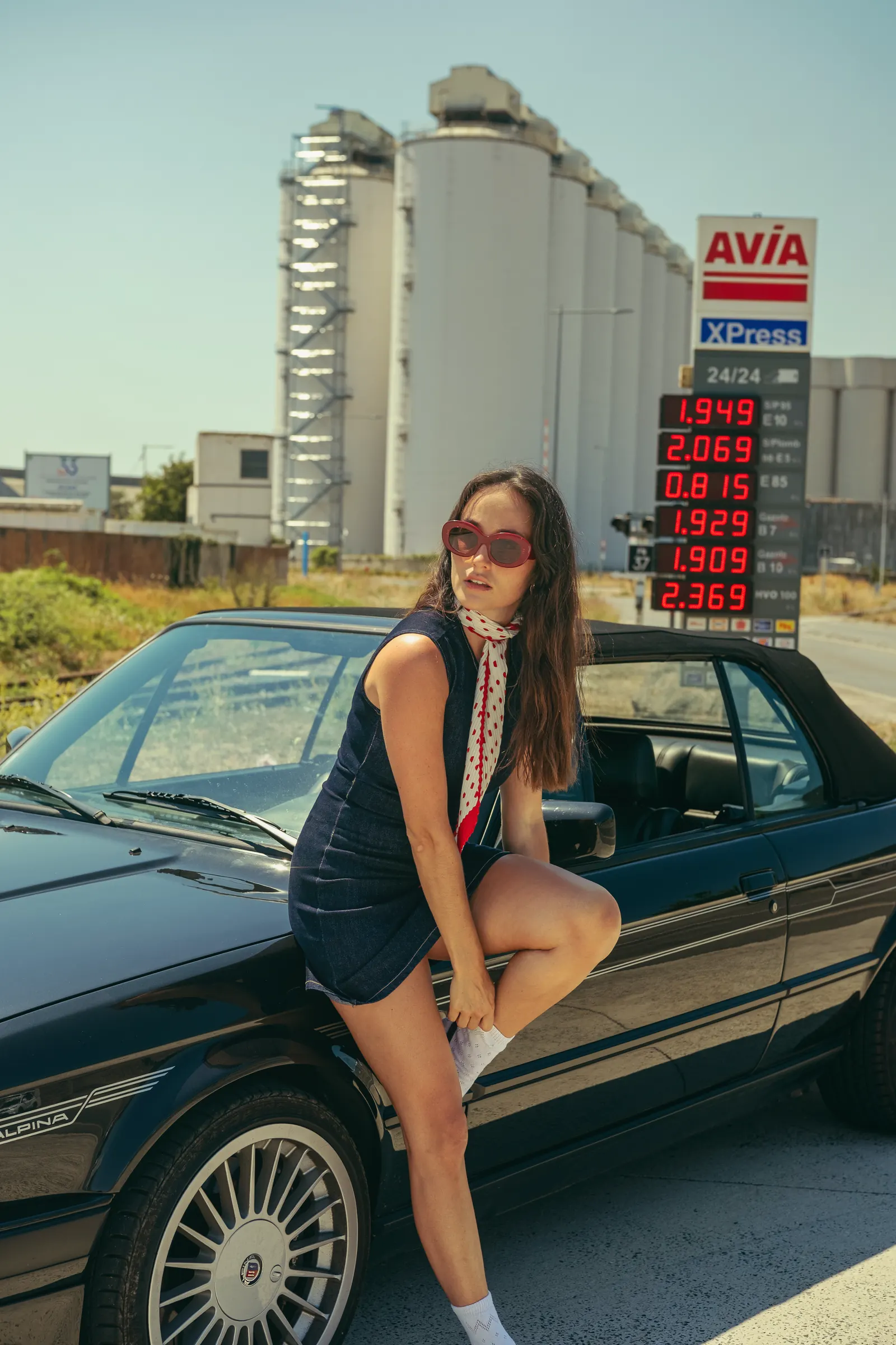 Femme en robe de jean et foulard à pois rouges, lunettes ovales rouges, appuyée sur un cabriolet noir devant les silos du port