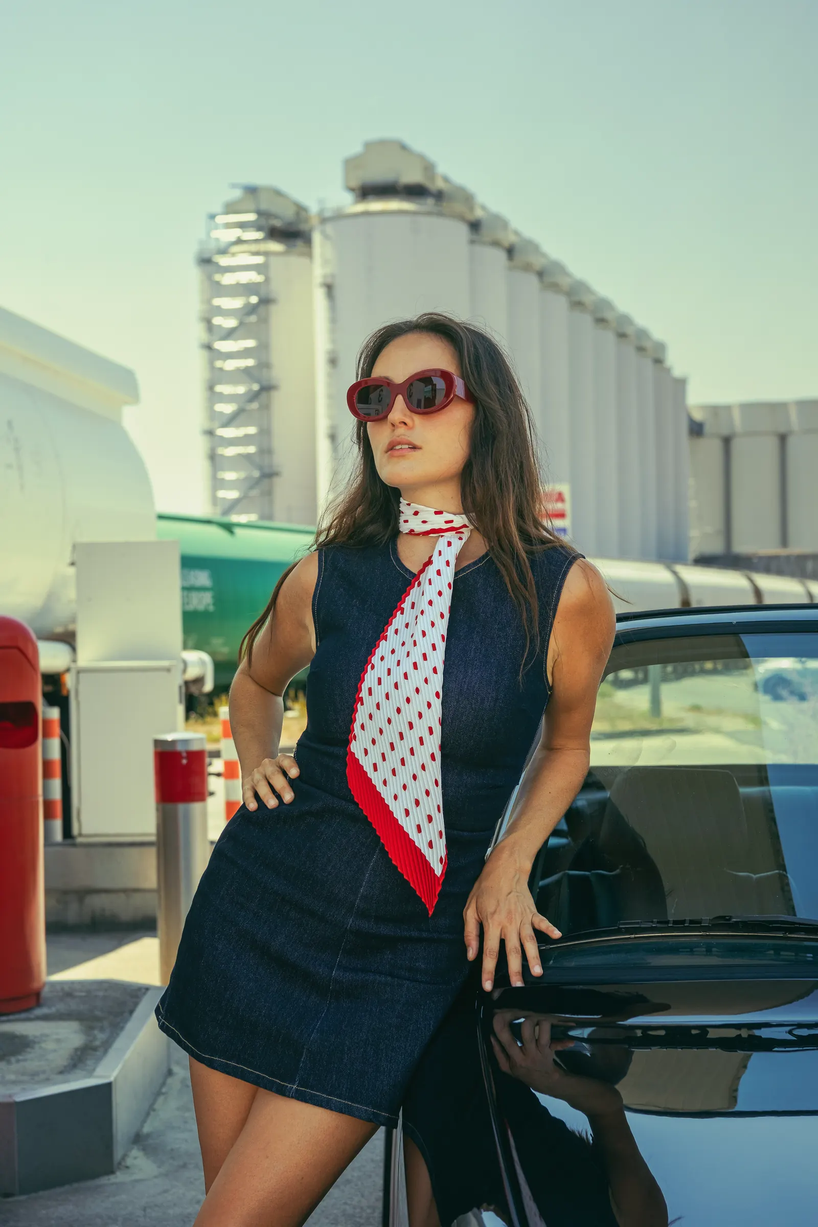 Femme en robe de jean sans manches et foulard blanc à pois rouges, lunettes ovales bordeaux, adossée à une décapotable noire