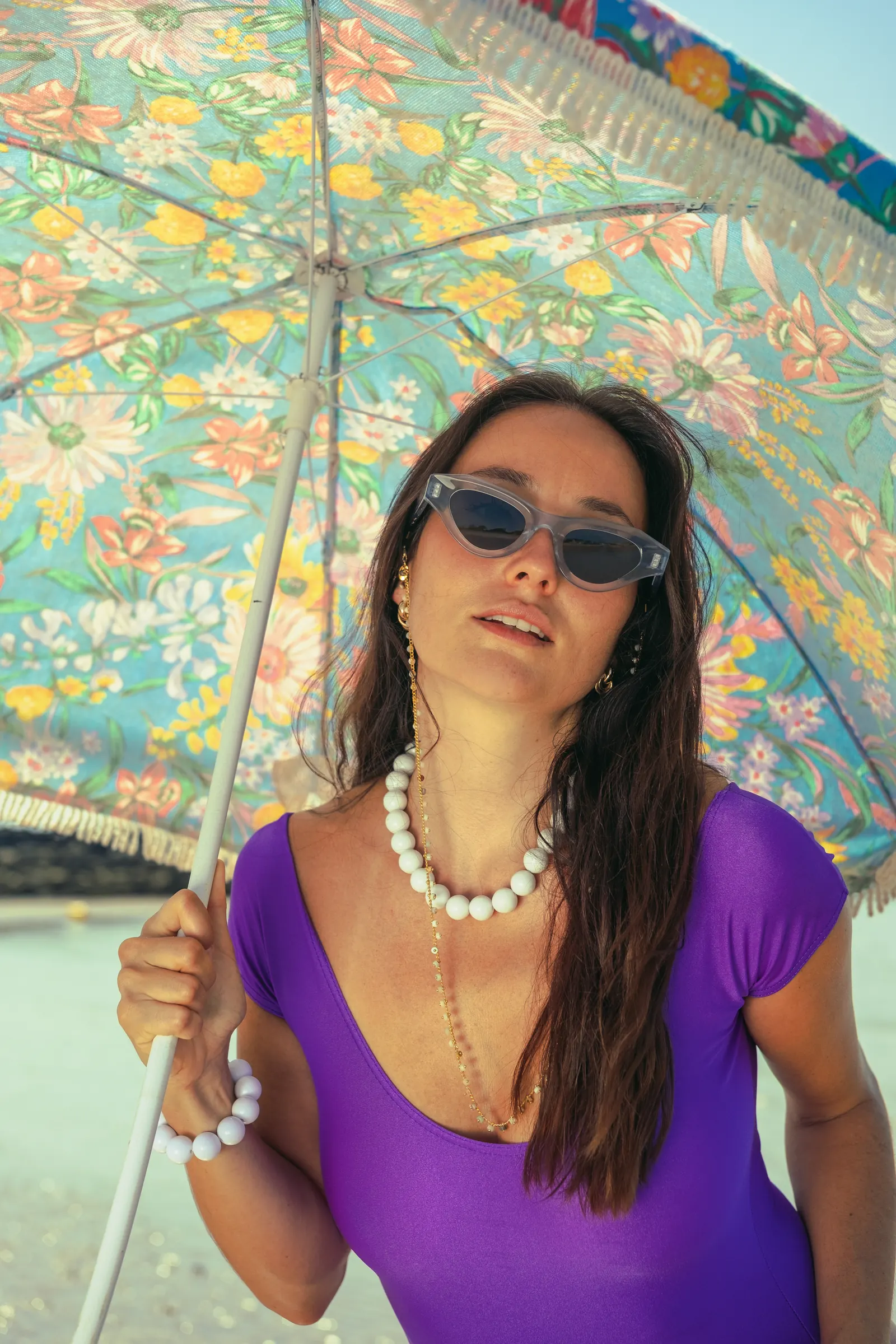 Portrait rapproché sous un parasol fleuri, lunettes cat-eye bleu givré avec chaînette, collier de perles blanches