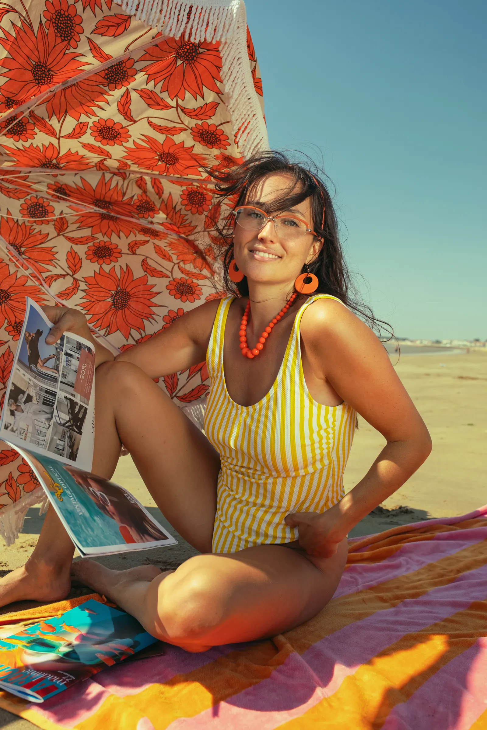 Femme en maillot rayé jaune assise sur une serviette, lisant un magazine Vintage Optical sous un parasol orange à fleurs