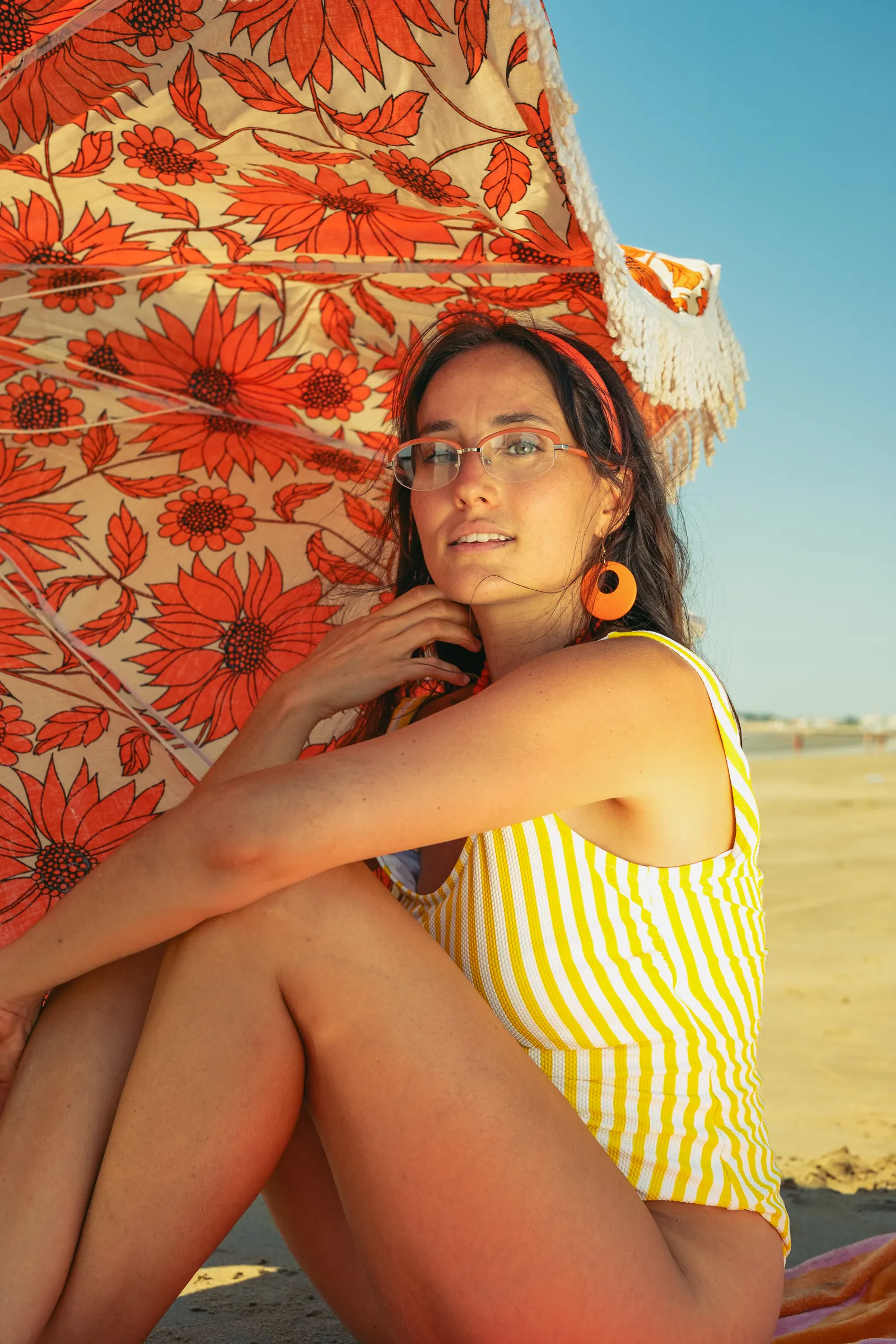 Femme assise genoux repliés sous un parasol orange, monture de vue fine orange, grandes boucles d’oreilles assorties